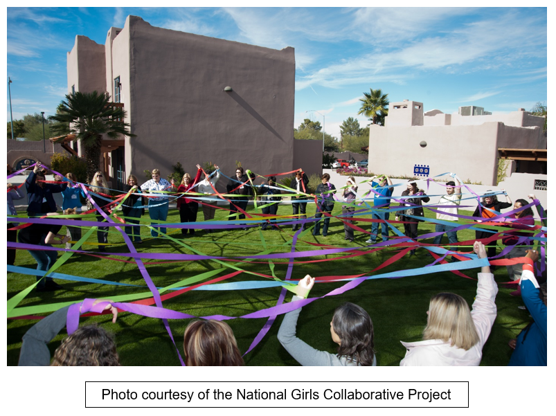 Women and girls gather in a large circle and hold ribbons. Text reads: 
Photo courtesy of the National Girls Collaborative project."