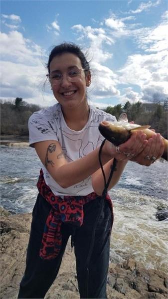 A young woman holds up a fish near a stream. 